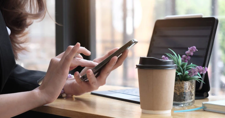 A person working at a cafe table, using a smartphone beside a laptop, takeaway coffee cup, and small potted plant, illustrating a quiet remote working environment.